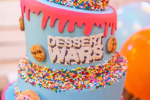 A colorful two-tier cake with blue frosting, sprinkles, cookie decorations, and a "Dessert Wars" sign.