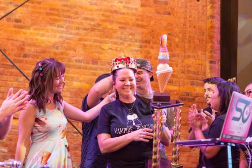 A woman wearing a crown smiles while holding an ice cream cone, surrounded by cheering friends in a festive setting.