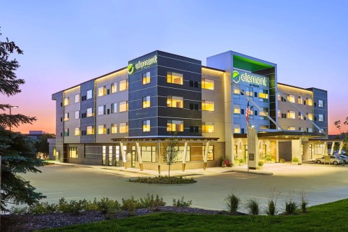 Modern hotel exterior at dusk, featuring large windows and a green logo, surrounded by landscaped grounds.
