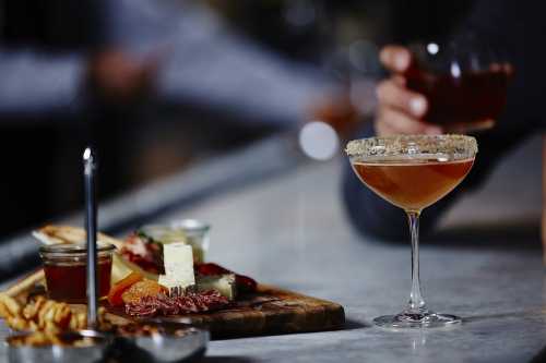 A cocktail with a sugared rim sits beside a charcuterie board featuring cheese, meats, and garnishes on a bar counter.