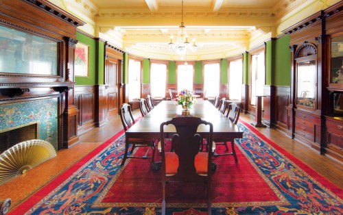 A spacious dining room with a long wooden table, elegant chairs, and ornate decor, featuring a colorful rug and large windows.