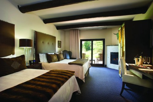 Cozy hotel room with two beds, a fireplace, and large windows overlooking a green outdoor area.