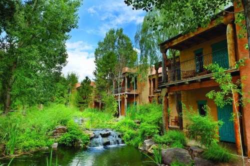 A serene landscape featuring a small waterfall, lush greenery, and adobe-style buildings under a blue sky.