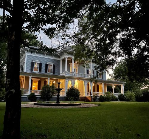 A large, elegant house with a porch, surrounded by trees and a manicured lawn, illuminated at dusk.