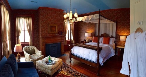 Cozy bedroom with a four-poster bed, vintage decor, a fireplace, and a seating area. Soft lighting enhances the ambiance.