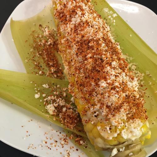 A plate of corn on the cob topped with cheese and spices, resting on green corn husks.