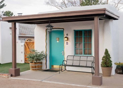 A charming adobe-style entrance with a turquoise door, bench, and potted plants under a wooden awning.