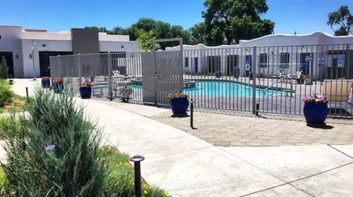 A fenced pool area with blue water, surrounded by potted plants and a pathway, under a clear blue sky.