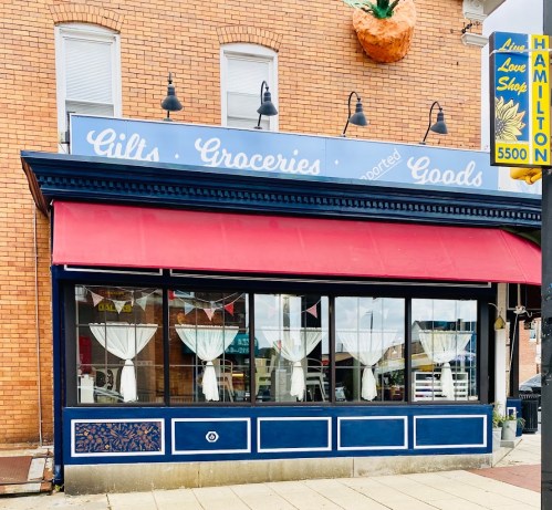 A storefront with a red awning, featuring signs for gifts, groceries, and goods, with white curtains in the windows.