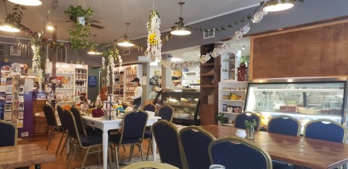 Cozy café interior with wooden tables, blue chairs, plants, and a display case filled with pastries.