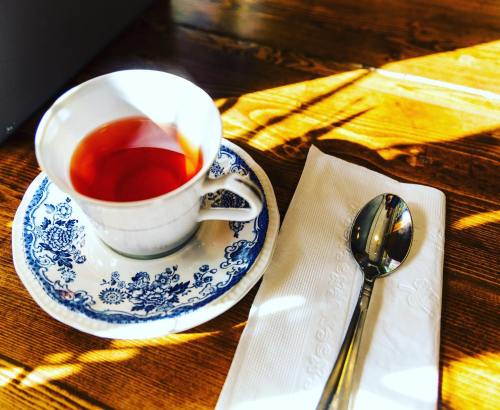 A cup of tea on a decorative saucer, accompanied by a spoon and a napkin, resting on a wooden table.