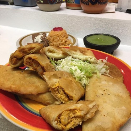 A colorful plate featuring crispy fried pastries filled with chicken, alongside rice, beans, and green salsa.