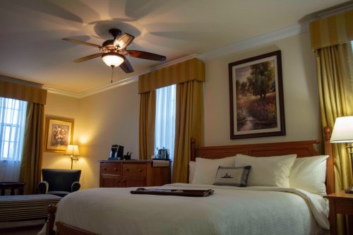 Cozy hotel room with a king bed, ceiling fan, and warm lighting, featuring a seating area and artwork on the walls.