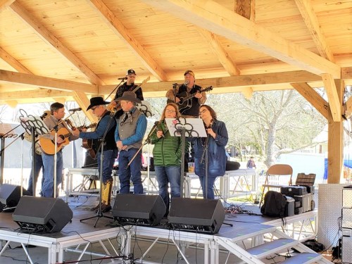 A group of musicians performs on stage under a wooden structure, playing instruments and singing together.