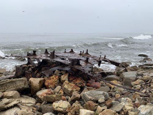 A weathered shipwreck rests on rocky shore, partially submerged in the ocean with waves gently lapping at it.