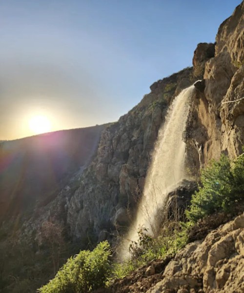 A waterfall cascades down rocky cliffs with a sunset in the background, illuminating the scene with warm light.