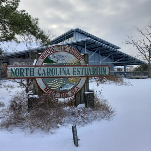 Sign for the North Carolina Estuarium, surrounded by snow, with a building in the background under a cloudy sky.