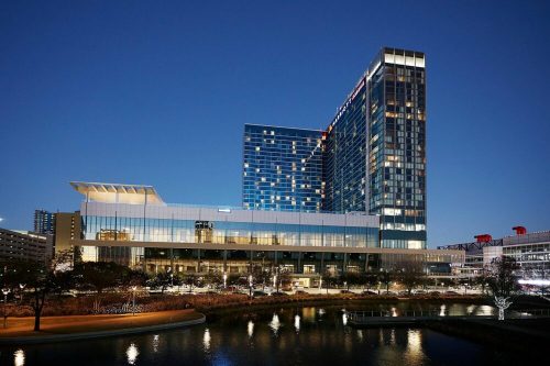 A modern hotel building illuminated at night, reflecting in a nearby water feature.