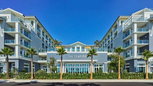 Modern hotel building with balconies, palm trees, and a clear blue sky. The entrance features large glass doors.