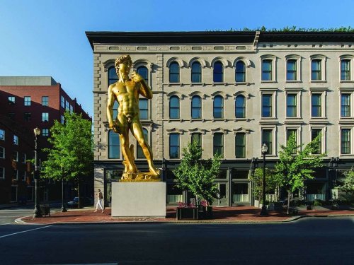 A large golden statue of a muscular figure stands in front of a historic building, surrounded by trees and a clear sky.