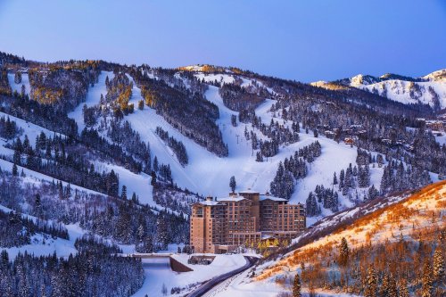 A snowy mountain landscape featuring a large hotel nestled among trees and ski slopes under a clear blue sky.