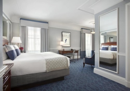 A stylish hotel room featuring a large bed, desk, mirror, and elegant decor with natural light from the windows.