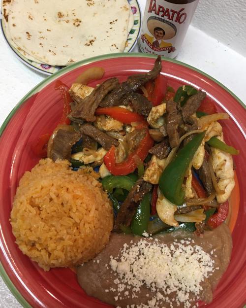 A colorful plate of fajitas with rice, refried beans topped with cheese, a tortilla, and a bottle of hot sauce.
