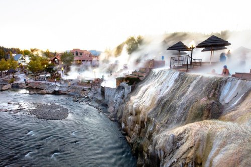 A scenic view of hot springs with steam rising, surrounded by trees and buildings along a riverbank at sunset.