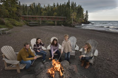 A family gathers around a campfire on a beach, roasting marshmallows with trees and a bridge in the background.