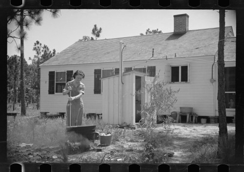 A woman in a polka dot dress cooks outdoors near a small white house surrounded by trees.