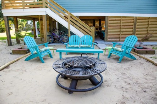 A cozy outdoor seating area with turquoise chairs, a fire pit, and bicycles in the background, set on sandy ground.