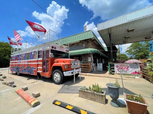 A colorful bus parked outside a rustic building with flags, surrounded by greenery and decorative planters.