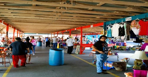 A bustling market scene with vendors and shoppers under a wooden structure, showcasing various goods and clothing.
