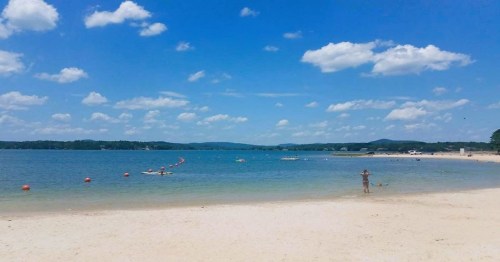A sunny beach scene with clear blue water, fluffy clouds, and a person wading near the shore.