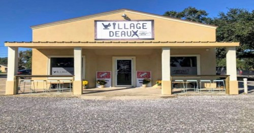 A small, yellow building with a sign reading "Village Deaux," featuring outdoor seating and flower pots.