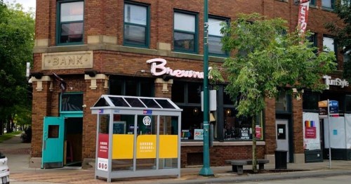 A brick building with a sign reading "Brewery" and a bus stop in front, featuring colorful advertisements.