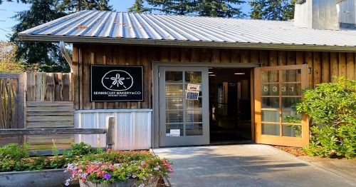 Exterior of a rustic bakery with a sign reading "Scrub Biscuit Bakery Co." and open doors, surrounded by greenery.