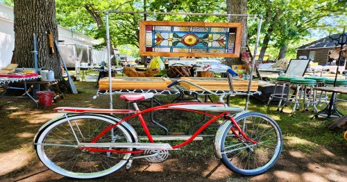 A vintage red bicycle stands beside a colorful stained glass piece in a sunny outdoor market setting.
