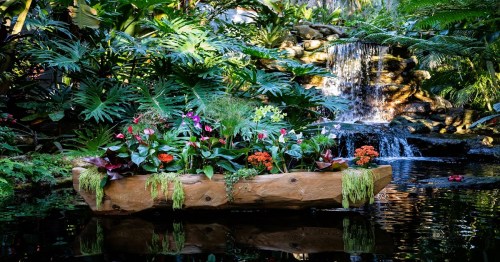 A serene garden scene featuring a wooden boat filled with colorful flowers, set against a waterfall and lush greenery.