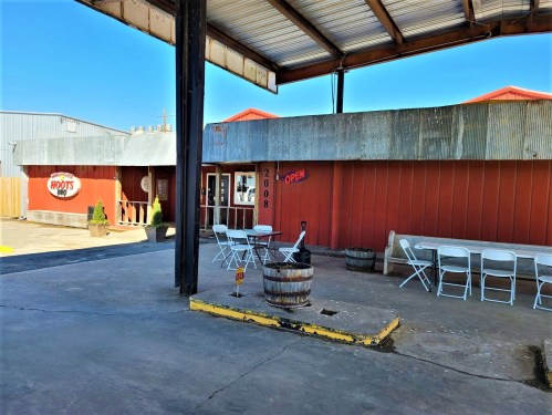 A rustic building with a sign reading "Open," featuring outdoor seating and a barrel, under a clear blue sky.