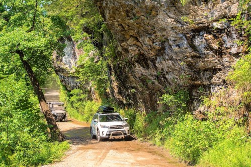 A white SUV drives along a narrow, muddy road surrounded by lush greenery and rocky cliffs.