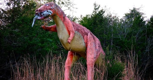 A colorful dinosaur statue stands in a grassy area, holding a smaller animal in its mouth, surrounded by trees.