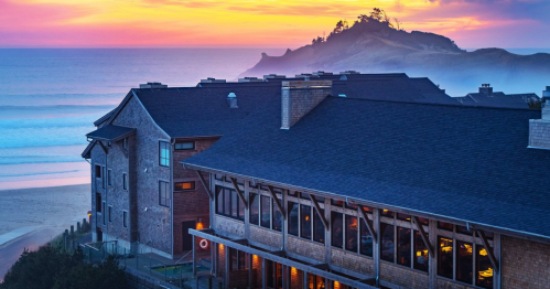 A coastal view at sunset, featuring a beachside building and a distant rocky island under a colorful sky.