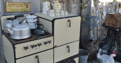 Vintage kitchen scene featuring an old stove, kettles, and decorative mugs against a rustic backdrop.