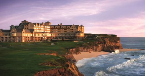 A coastal hotel sits atop a cliff, overlooking the ocean at sunset, with waves crashing on the beach below.