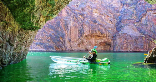 A person kayaking in a serene green lake surrounded by rocky cliffs and a cave entrance.