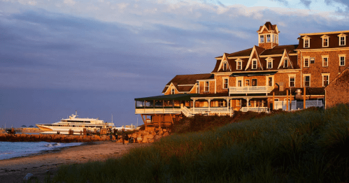 A coastal hotel at sunset, with a boat docked nearby and grassy dunes in the foreground.