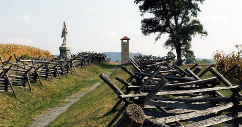 A pathway lined with wooden fences, leading to a tower and a statue, surrounded by fields and trees.