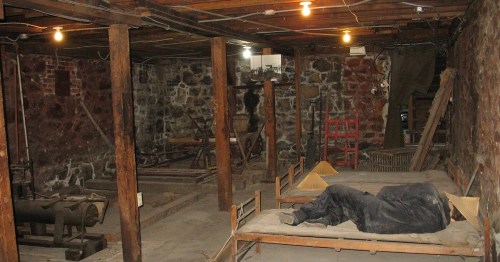 A dimly lit basement with stone walls, wooden beams, and two beds; one bed is occupied by a person lying down.