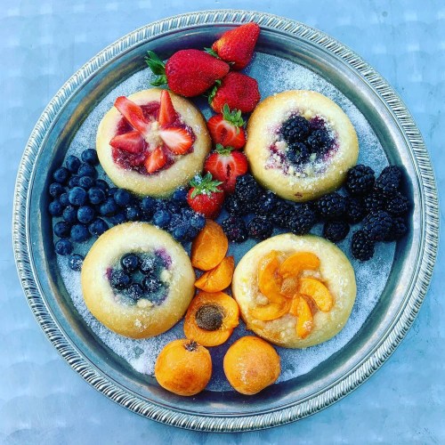 A silver platter with four fruit-topped pastries surrounded by fresh strawberries, blueberries, blackberries, and apricots.